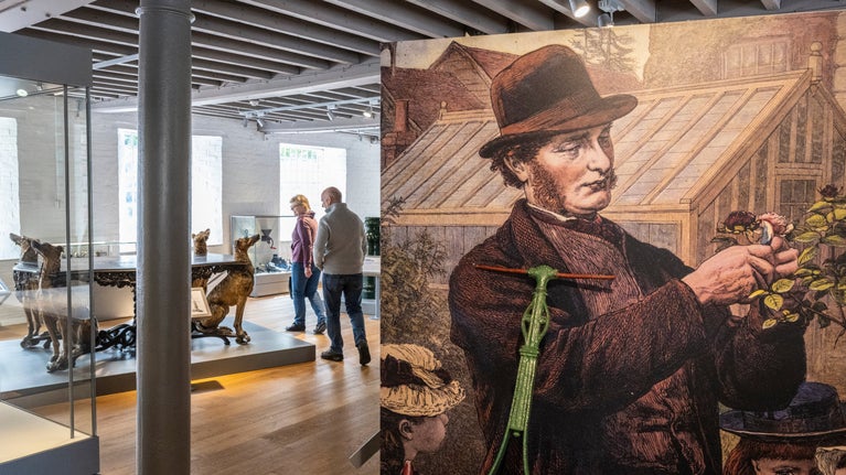 Two visitors among the exhibits at the Museum of Iron. They are walking towards a large iron table with iron dogs for legs. A drawing of a man in Victorian attire is on a board nearest the camera on the right.